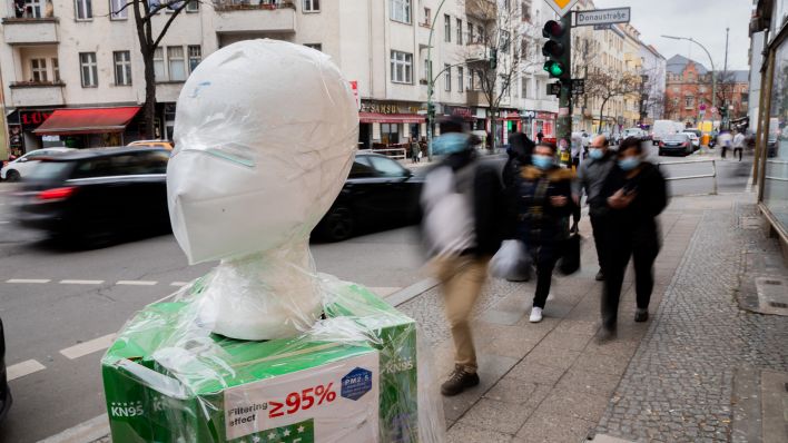 Eine FFP2-Maske wird vor einem Geschäft in Berlin-Neukölln ausgestellt, wobei Passanten vorbeigehen. (Quelle: dpa/Christoph Soeder)