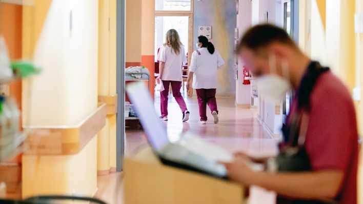 Zwei Mitarbeiterinnen gehen durch den Flur eines Krankenhauses, wobei ein Krankenpfleger an einem mobilen Computerarbeitsplatz steht. (Quelle: dpa/Christoph Soeder)