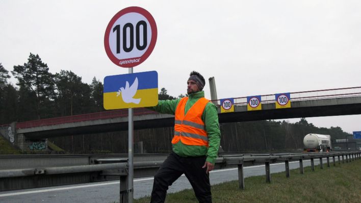 Ein Demonstrant von Greenpeace hält an der Autobahn ein Schild mit der ukrainischen Flagge, auf die eine Friedenstaube gemalt ist, und einem Schild für eine Tempobegrenzung auf 100. (Quelle: dpa/Paul Zinken)