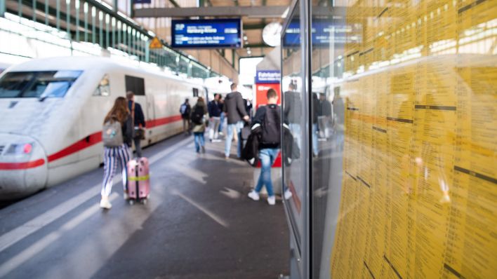 Ein Fahrplan hängt an einem Bahnhof. Im Hintergrund gehen Reisende zu einem ICE. (Quelle: dpa/Tom Weller)
