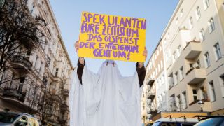 Eine Aktivistin im Gespensterkostüm hält ein Protestschild nahe dem Organisationsbüro des Volksbegehrens «Deutsche Wohnen & Co. enteignen» in die Kamera. (Quelle: dpa/Christoph Soeder)
