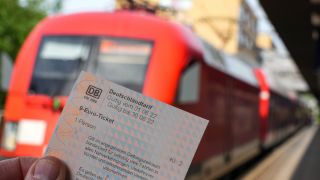 Die Hand eines Mannes hält auf dem Potsdamer Hauptbahnhof ein 9-Euro-Ticket. Im Hintergrund fährt ein Regionalzug der Bahn nach Frankfurt/Oder. (Quelle: Soeren Stache/dpa)