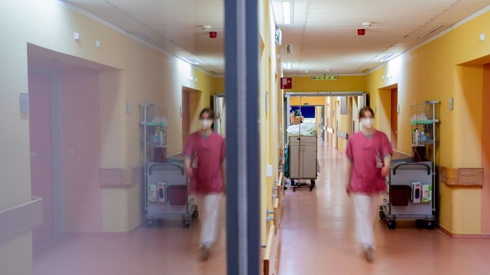 Eine Pflegehelferin geht im Gang eines Krankenhauses, wobei sie sich in einer Glastür spiegelt. (Quelle: dpa/Christoph Soeder)