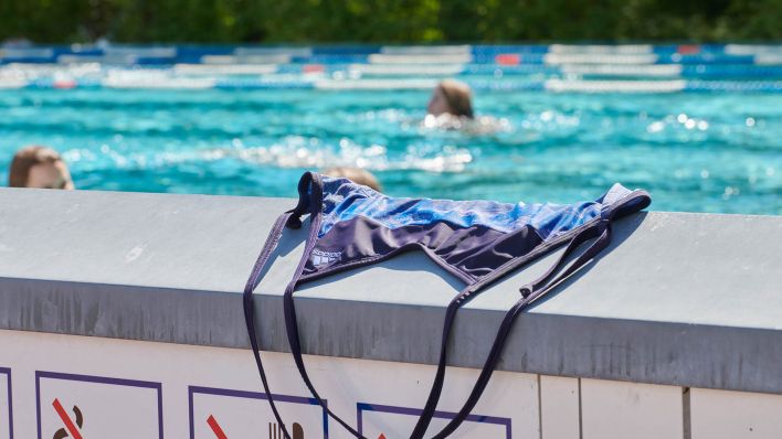 Symbolbild: Ein Badeanzug liegt auf einer Mauer im Prinzenbad, dahinter schwimmen Menschen im Becken ihre Bahnen. (Quelle: dpa/A. Riedl)