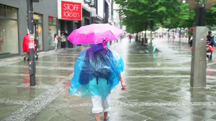 Eine Frau läuft mit Regenschirm und Regencape durch eine Einkaufsstraße. (Quelle:rbb)