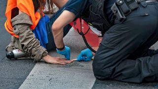 Archivbild: Ein Polizeibeamter löst am 01.07.2022 auf einer Kreuzung am Kaiserdamm die auf der Straße klebende Hand eines Demonstranten der Gruppe "Letzte Generation". (Quelle: dpa/Paul Zinken)