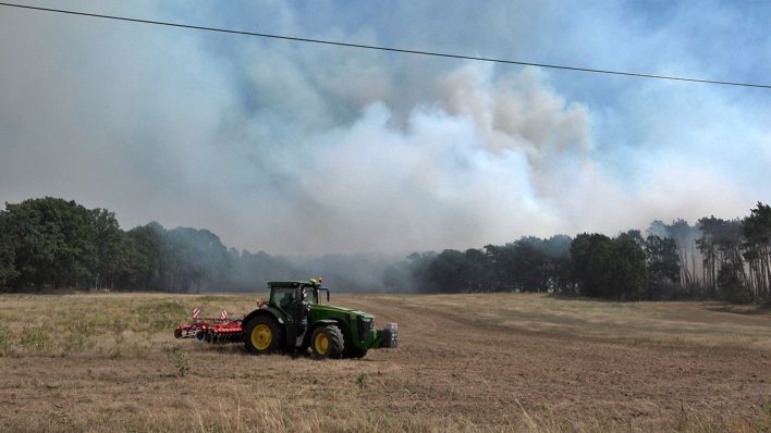 Ein Traktor steht am 25.07.2022 auf einem Feld vor einem Waldstück bei Gransee, aus dem Rauchwolken aufsteigen. (Quelle: dpa/Christian Guttmann)