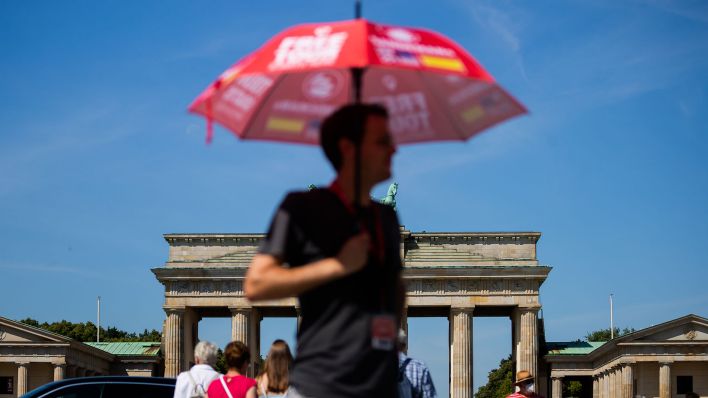 Ein Mann steht vor dem Brandenburger Tor und schützt sich mit einem Regenschirm vor der Sonne. (Quelle: dpa/Christoph Soeder).