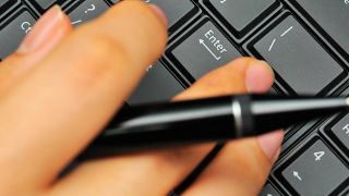 Eine Hand hält einen Stift über einer Computertastatur. (Quelle: imago/YAY Images)