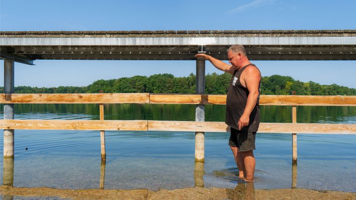 Der Bootsverleiher Mario Schmitz demonstriert mit seiner Körperhöhe am 20.07.2022 das Absinken des Wasserpegels des Strausberger Sees (Quelle: rbb/Hinze).