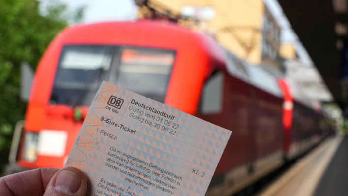 Die Hand eines Mannes hält auf dem Potsdamer Hauptbahnhof ein 9-Euro-Ticket. Im Hintergrund fährt ein Regionalzug der Bahn nach Frankfurt/Oder. (Foto: Soeren Stache/dpa )