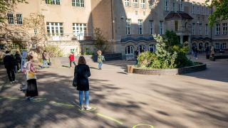 Symbolbild: Schüler*innen stehen vor einer Schule auf dem Schulhof. (Quelle: dpa/K. Nietfeld)