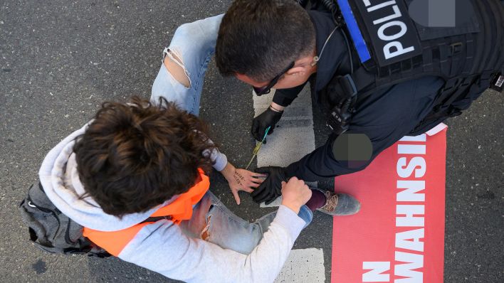 Archivbild: Umweltaktivisten der Gruppe «Letzte Generation» haben sich auf der Fahrbahn fest geklebt und ein Polizist versucht den Kleber zu lösen. (Quelle: dpa/R. Michael)