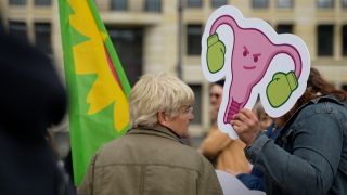 Eine Person hält ein Pappschild von einem Uterus mit Boxhandschuhen in die Luft. Der Uterus hat ein Gesicht und guckt fies. Im Vordergrund steht eine Frau, mit einer Fahne der Partei Bündnis 90/Die Grünen.