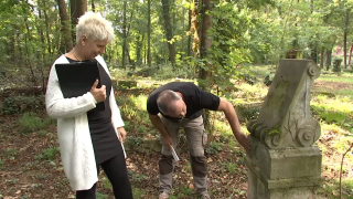 Ein Mann bückt sich zu einem antiken Grabstein hinunter. Eine Frau, die ein Klemmbrett hält, schaut ihm dabei zu.