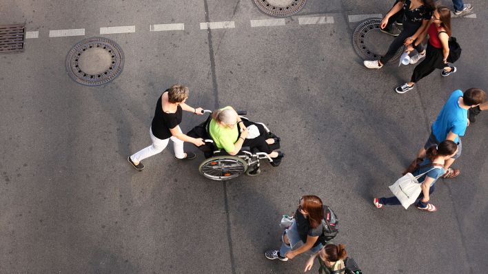 Symbolfoto: Eine Frau schiebt am 16.09.2018 eine Seniorin im Rollstuhl auf einer Einkaufsstraße in Berlin-Friedenau über eine Ampelkreuzung. Die alte Dame hält dabei eine Tüte in ihrem Schoß (Quelle: dpa / Wolfram Steinberg).
