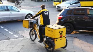 Ein Briefbote von Deutsche Post DHL steht mit seinem Elektrofahrrad an einer Straße in Berlin. (Quelle: dpa/Wolfram Steinberg)