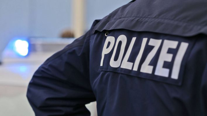 Polizei-Schriftzug auf dem Ruecken eines Polizisten mit Blaulich im Hintergrund. (Quelle: imago images)