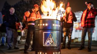 Symbolbild: Beschäftigte bei einem Warnstreik hinter einem Feuerofen, der das Logo der Gewerkschaft IG Metall zeigt. (Quelle: dpa/F.Rumpenhorst)