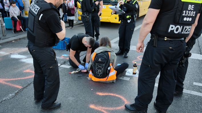 Symbolbild: Ein Polizist versucht die festgeklebte Hand eines Mannes vom Asphalt zu lösen, während Polizisten versuchen eine Blockade der Protestgruppe „letzte Generation“ vor dem Berliner Hauptbahnhof auf der Invalidenstraße aufzulösen