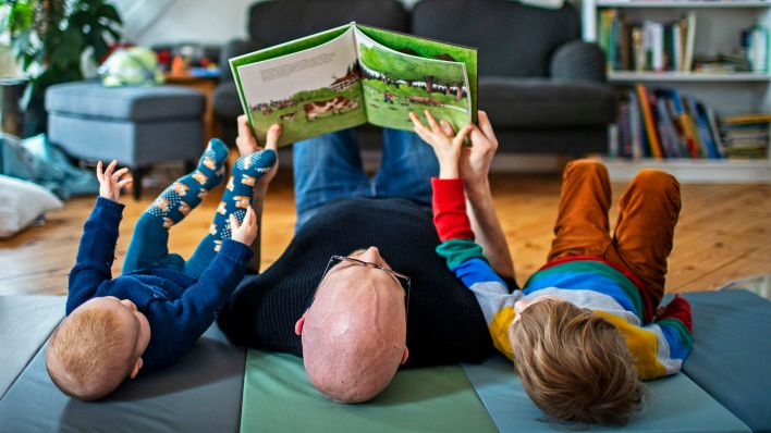 Symbolfoto: Ein Kinderbetreuer liest mit seinen beiden Kindern auf dem Boden liegend ein Buch.(Quelle:dpa/J.Schmitz)