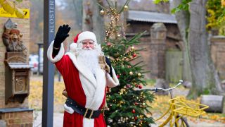 Archivbild:Der Weihnachtsmann winkt am 11.11.2021 neben dem Haus, vor einem Weihnachtsbaum und seinem Rad in Himmelpfort.(Quelle:dpa/S.Stache)