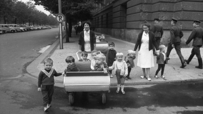 Archivbild: DDR-Krippenwagen mit Kindern, Erzieherinnen und Soldaten im Hintergrund. (Quelle: dpa/K. Rose)