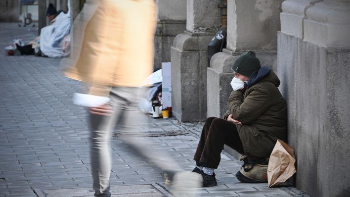 Archivbild: Ein Obdachloser Mann sitzt mit Maske auf der Straße, an einem Gebäude angelehnt. (Quelle: dpa/S. Simon)