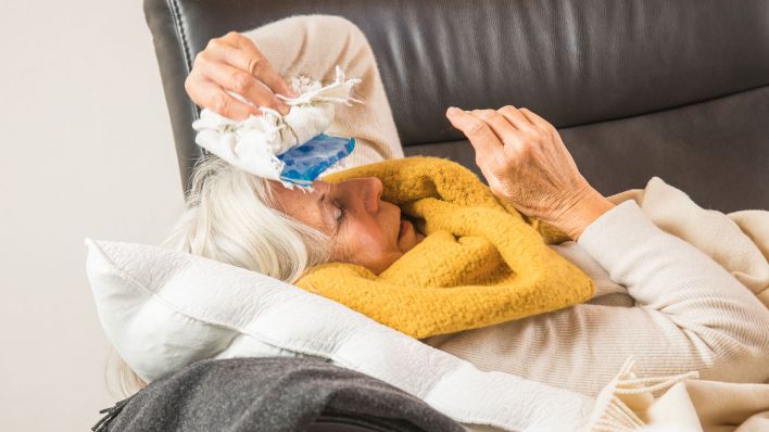 Eine ältere Frau liegt krank auf ihrer Couch und kühlt ihre Stirn. (Quelle: dpa/Christin Klose)