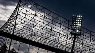 Symbolbild: Flutlichtanlage in einem Fußballstadion ist durch das Netz eines Fußballtors zu sehen. (Quelle: dpa/J. Ferreira)