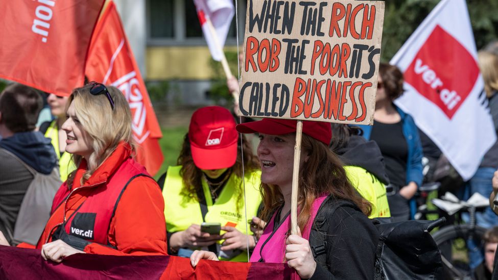 Eine Teilnehmerin hält am 1.05.2023 ein Schild mit der Aufschrift „When the rich rob the poor it’s called business“.(Quelle:dpa/H.P.Albert)