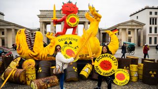 Touristen von den Philippinen posieren mit Schwertern und Schildern vor einer Dinosaurier-Figur und der roten Sonne, teil des Anti-Atomkraft-Logos, die bei einer Aktion der Umweltorganisation Greenpeace vor dem Brandenburger Tor liegt. (Quelle: dpa/C. Soeder)