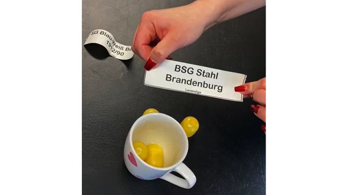 Eine Hand hält ein Los mit der Aufschrift: "BSG Stahl Brandenburg Landesliga":(Quelle:Screenshot aus Video)