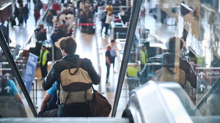 Ein Mann geht am 02.10.2022 eine Rolltreppe hinunter, während Personen am Flughafen Berlin Brandenburg (BER) "Willy Brandt" an Check-in-Schaltern warten. (Quelle: dpa-Bildfunk/Annette Riedl)