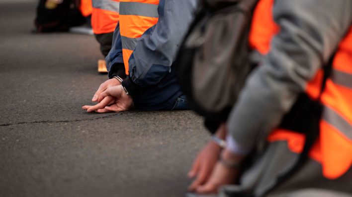 Symbolbild:Aktivisten der Gruppierung Letzte Generation sitzen mit Handschellen, die ihnen von der Polizei angelegt wurden, auf einer STraße.(Quelle:dpa/P.Zinken)
