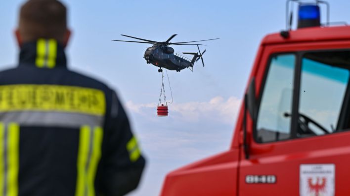 Ein Hubschrauber der Luftwaffe fliegt mit einem Wasserbehälter während einer Übung über den Flugplatz. (Quelle: dpa/Patrick Pleul)