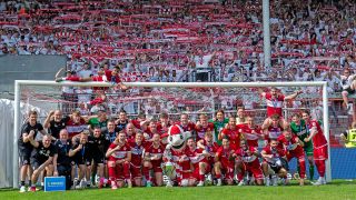 Die Mannschaft von Energie Cottbus posiert mit dem Meisterpokal und den Fans im Rücken (Quelle: IMAGO/Fotostand)