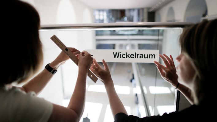 Symbolbild: Helferinnen kleben in Berlin ein Hinweisschild mit der Aufschrift "Wickelraum" an ein Geländer. (Quelle: dpa/K. Nietfeld)