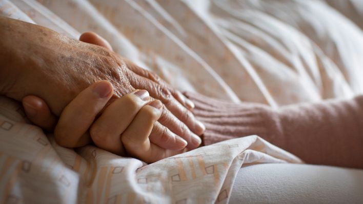 Symbolbild:Eine Hand einer im Bett liegenden Person wird von einer anderen Hand gehalten.(Quelle:picture alliance/U.Grabowsky)