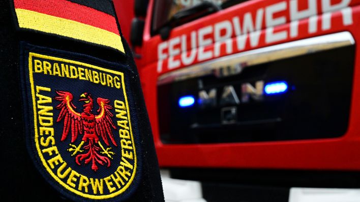 Symbolbild: Ein Abzeichen "Landesfeuerwehrverband Brandenburg" ist am Ärmel eines Feuerwehrmannes neben einem Feuerwehrfahrzeug zu sehen