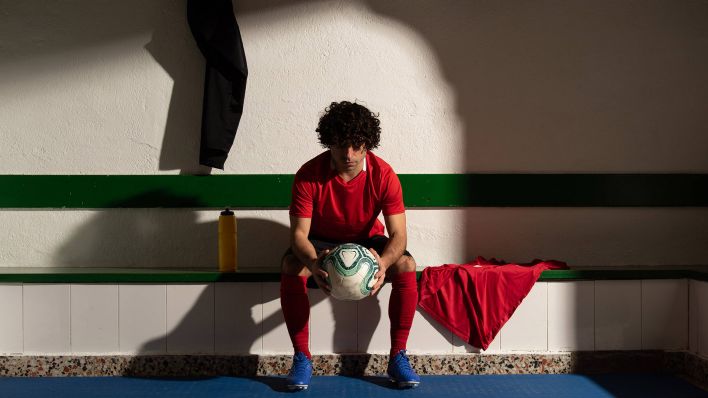 Symbolbild: Ein Fußballspieler sitzt in der Umkleidekabine im Halbdunkeln und hält nachdenklich einen Fußball in den Händen. (Quelle: imago images/E. Bengoetxea)