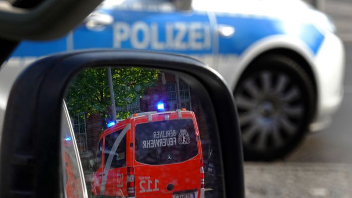 sYMBOLBILD. Notarztwagen der Berliner Feuerwehr spiegelt sich im Seitenspiegel eines PKW. Ein Einsatzfahrzeug der Polizei steht davor. (Quelle: IMAGO/Frank Sorge)