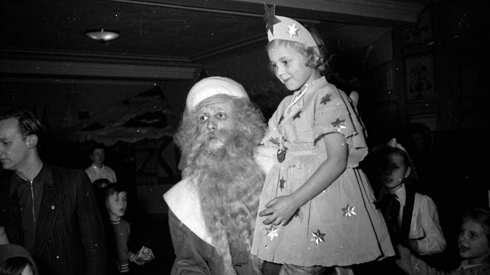 Die Kinderweihnachtsfeier des Werks für Fernmeldewesen in Berlin-Oberschöneweide 1954 im WF-Kulturhaus: Ein Weihnachtsmann trägt ein Mädchen in einem Sternenkostüm. Daneben stehen ein paar Kinder und Erwachsene (Quelle: Industriesalon Schöneweide).