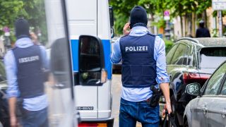Symbolbild:Ein Polizist steht bei einer Hausdurchsuchung neben einem Fahrzeug.(Quelle:dpa/C.Soeder)