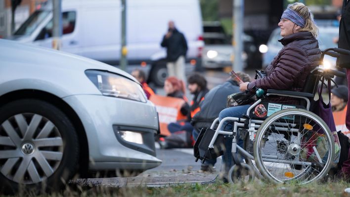 Eine Frau im Rollstuhl ist während einer Straßenblockade der Klimaschutzgruppe "Letzte Generation" an einer Abfahrt der A10 (Quelle: dpa/