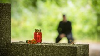 Symbolbild: Eine Grabkerze steht am 25.06.2021 in Berlin auf einem Grabstein, waehrend im Hintergrund ein Mann auf einer Bank sitzt.(Quelle:dpa/C.Klose)
