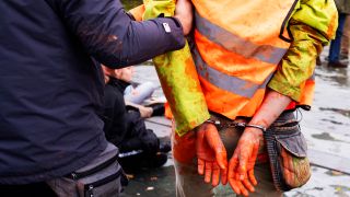 Symbolbild:Ein Polizist führt einen mit orangener Farbe beschmierten Aktivisten in Handschellen weg.(Quelle:picture alliance/AP/M.Schreiber)