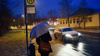 Symbolbild: Eine Frau steht in Brandenburg während des ersten Schneefalls mit einem Regenschirm an einer Bushaltestelle. (Quelle: dpa/Fischer)