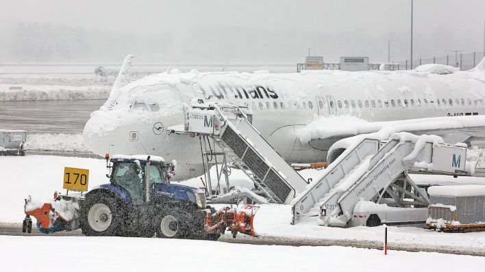 Ein Räum- und Streufahrzeug fährt am 02.12.2023 im Schneetreiben auf dem Flughafen München an einer verschneiten Maschine von Lufthansa vorbei. (Quelle: dpa/Karl-Josef Hildenbrand)