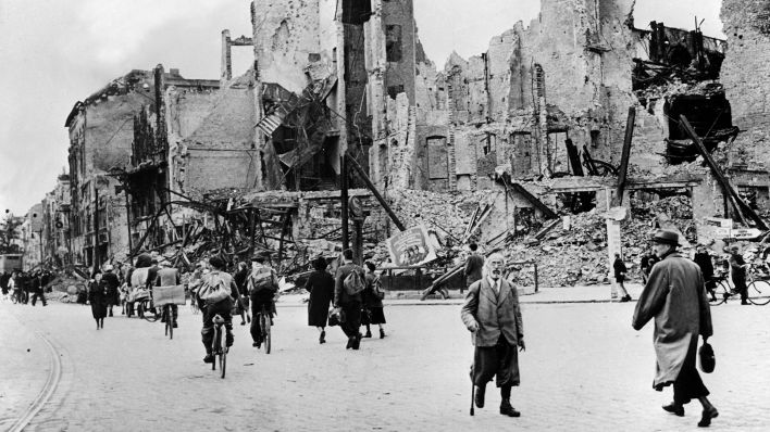 Menschen gehen nach Kriegsende im Mai 1945 auf einer Straße an zerstörten Häusern entlang. (Quelle: dpa-Bildfunk)